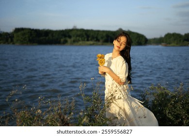 Serene Woman in White Dress Holding Flowers by a Tranquil Lakeside at Sunset - Peaceful Nature and Outdoor Beauty Scene. - Powered by Shutterstock