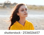 Serene woman in yellow hoodie standing on sandy beach with closed eyes gazing towards the sky