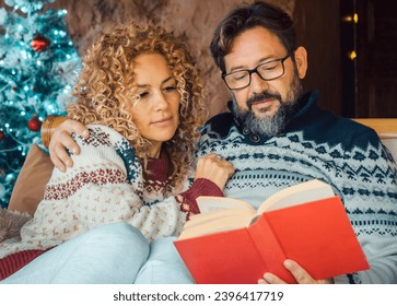 Serene young mature couple at home enjoying relax and indoor leisure activity in december holiday. Christmas tree in background. Man reading a book with a woman. Married together relationship love - Shutterstock ID 2396417719