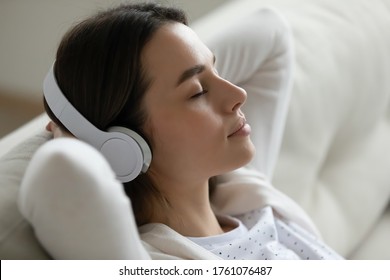 Serene Young Woman Closed Eyes Relaxing Leaned On Couch Puts Hands Behind Head Listens Meditative Music Through Modern Wireless Headphones, Close Up View. Free Time, No Stress, Lounge At Home Concept