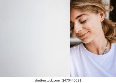 Serene young woman leaning against a white wall with eyes closed, exuding relaxation and peace. - Powered by Shutterstock