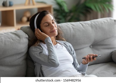 Serene young woman resting on sofa wearing casual home clothes wireless headphones enjoy weekend free time using online application on cellphone listens music, closed eyes reduces anxiety and fatigue - Powered by Shutterstock