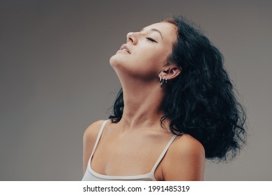 Serene young woman taking a quiet moment to relax with head tilted back, parted lips and closed eyes in a head and shoulders portrait against a grey studio background