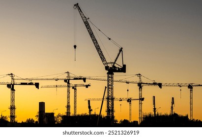 A series of construction cranes rise against a colorful sunset, casting long shadows over a busy construction site, signaling activity in the early evening light. - Powered by Shutterstock