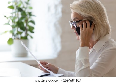 Serious 60 years old businesswoman talking on smartphone and reading document, solving corporate business problem. Female employee discussing document by cellphone, receive news. - Powered by Shutterstock