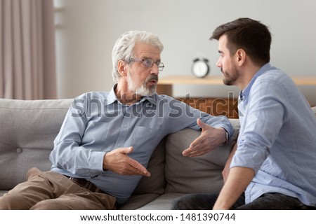 Serious 60s elderly father and grown up adult son sitting on sofa talking having important conversation trying to solve life issues problem, different men relative people communication at home concept