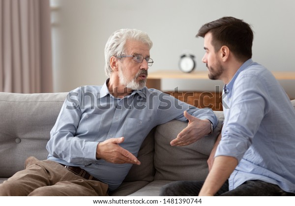 Serious 60s elderly father and grown up adult son\
sitting on sofa talking having important conversation trying to\
solve life issues problem, different men relative people\
communication at home\
concept