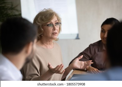 Serious 60s Mature Female Company Leader In Eyewear Holding Negotiations Meeting With Interested Diverse Young Colleagues, Sitting Together At Table, Discussing Working Issues Or Project Details.