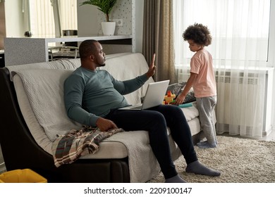 Serious Adult Father Showing Stop Gesture With Hand To His Little Son Playing With Construction Building Blocks At Home. Man Working On Laptop On Sofa. Young Black Family Lifestyle And Relationship