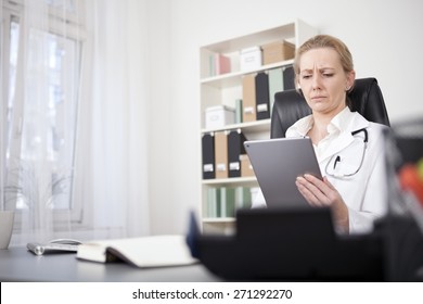 Serious Adult Female Medical Doctor Reading At Her Tablet Computer While Sitting At Her Desk.