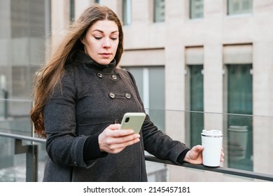 Serious Adult Female In Warm Clothes Using Cellphone While Standing Near Glass Fence With Tumbler Cup For Takeaway Hot Drink Against Tower