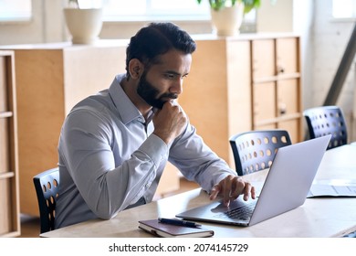 Serious adult thoughtful focused Indian Hispanic boss ceo businessman with hand on chin using typing on computer pc laptop working in contemporary office, accounting analysing report financial data. - Powered by Shutterstock