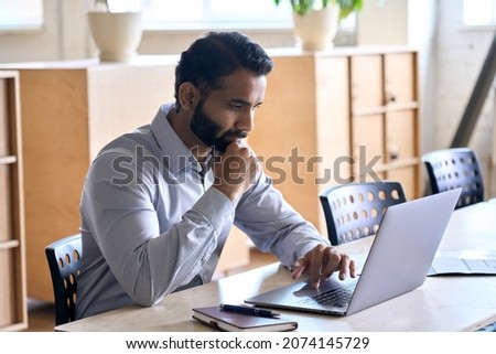 Serious adult thoughtful focused Indian Hispanic boss ceo businessman with hand on chin using typing on computer pc laptop working in contemporary office, accounting analysing report financial data.