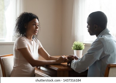 Serious african 30s married couple in love sitting at table having heart-to-heart intimate straight talk, mixed race wife holding hands of beloved black husband family share problems thoughts at home