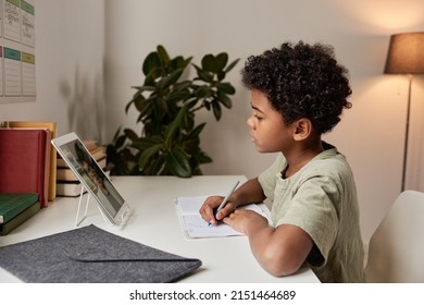 Serious African American boy with curly hair sitting at table with workbook and looking at tablet screen while attentively listening to teacher online
