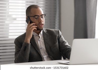 Serious African American businessman in glasses talking on phone, using laptop, having business conversation, hr manager holding interview by cellphone, employee consulting client, making sell offer