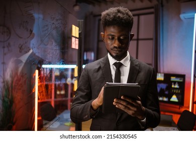 Serious African American Businessman Managing Project On Digital Tablet Pc, Working In Night Office With Scrum Glass Board. Focused Male Professional Planning Corporate Strategy Using Computer