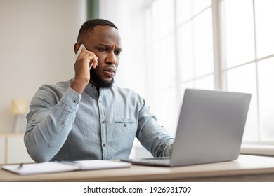 Serious African American Businessman Talking On Phone Using Laptop Sitting In Modern Office. Business Communication Concept. Entrepreneur Having Negotiations Via Cellphone Call