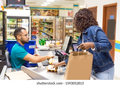 Serious African American Cashier Scanning Goods At Checkout. Back View Of Woman Packing Products In Paper Bag. Shopping Concept