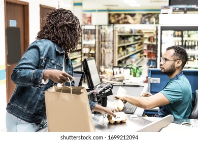 Serious African American Cashier Scanning Goods At Checkout. Back View Of Woman Packing Products In Paper Bag. Shopping Concept