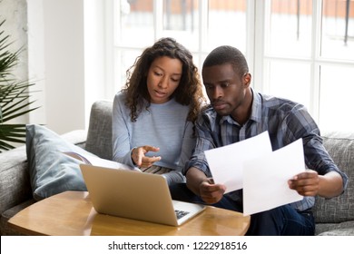 Serious African American couple discussing paper documents, sitting together on couch at home, man and woman checking bills, bank account balance, terms of contract, mortgage, loan agreement