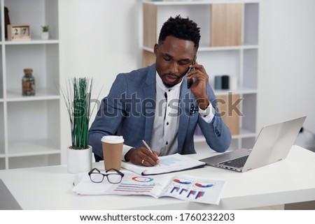 Serious african american entrepreneur has a phone conversation with business partners at cozy office, using laptop and makes some notes. Free copy space.