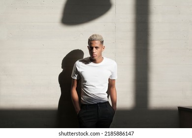 Serious African American Male In Casual Clothes Looking At Camera While Standing Against Wall With Sunlight And Shadow In City