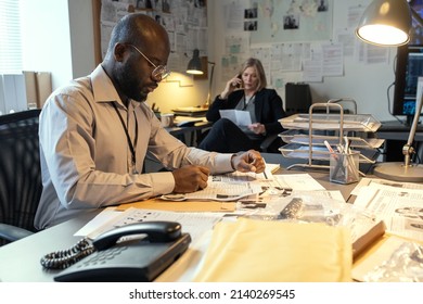 Serious African American Male Detective Making Notes About Criminal In His Profile While Sitting By Desk Against Mature Female Coworker