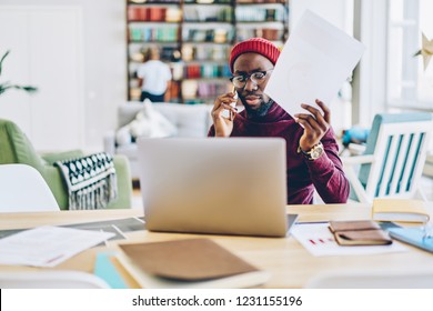 Serious African American Male Freelancer Concentrated On Remote Job At Home Holding Papers And Making Phone Call At Desktop, Pensive Dark Skinned Man Concentrated On Reading Online News On Netbook
