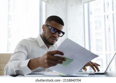 Serious African American Manager In Stylish Glasses Reading Corporate Document With Note, Sales Report, Boss Order, Doing Paperwork. Legal Expert Checking Document At Workplace With Laptop