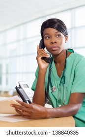 Serious African American Nurse At Hospital Work Station On The Phone With A Patient File And Stethoscope.