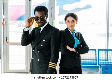 Serious African American Pilot In Sunglasses And Stewardess With Crossed Arms Standing Together In Airport