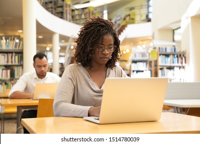 Serious African American Student Working On Research Paper In Library. People Sitting At Desks And Using Laptops In Computer Class With Bookshelves. Academic Research Concept