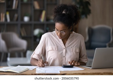 Serious African American woman in glasses calculating expenses, using calculator, sitting at desk with laptop and financial documents, focused young female managing planning household budget