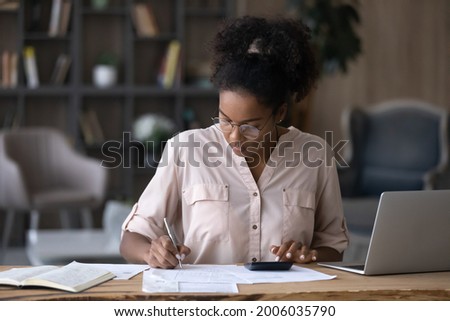 Serious African American woman in glasses calculating expenses, using calculator, sitting at desk with laptop and financial documents, focused young female managing planning household budget