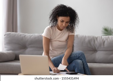 Serious African American woman using laptop, calculating finance at home, checking bills, focused girl using calculator, freelancer economist or financier writing report, student doing homework