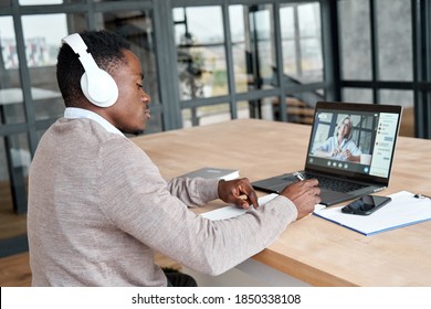 Serious African Business Man Or Black Male Student Wearing Headphones Conference Video Calling, Watching Webinar Online, Social Distance Learning Or Working Using Laptop At Home Office, Taking Notes.