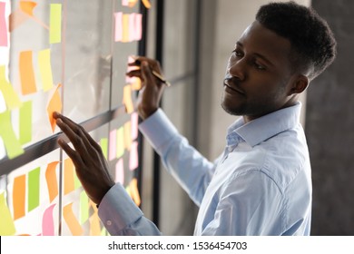 Serious african businessmen managing project tasks on sticky notes, focused male professional coach teacher write creative business idea on post it stickers planning corporate strategy on glass board
