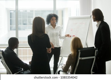 Serious african female leader or coach gives presentation of new marketing plan at sales team meeting, black employee explains colleagues new client management strategy idea at group office training