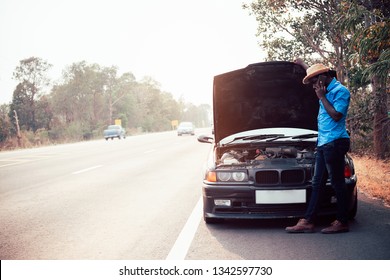 Serious african man using smartphone and holding his head by hands standing near his old broken car  with raised hood on the highway road - Powered by Shutterstock