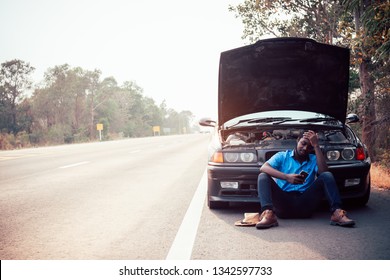 Serious African Man Using Smartphone And Holding His Head By Hands Standing Near His Old Broken Car  With Raised Hood On The Highway Road