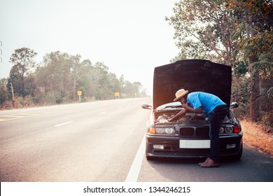 Serious African Man Using Smartphone And Holding His Head By Hands Standing Near His Old Broken Car  With Raised Hood On The Highway Road