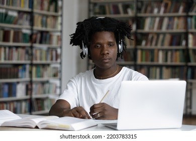 Serious African Student Guy Sit At Table Look At Laptop Makes Assignment, Listen Audio Through Headphones, Writes In Copybook. Prepare For University Exam, Study In Library Use Tech, Education Concept