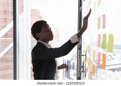 Serious African woman prepare boardroom for corporate briefing, prepare tasks attaching notes on glass wall. Start up ideas, create strategy development, makes schedule or agenda, to-do list concept