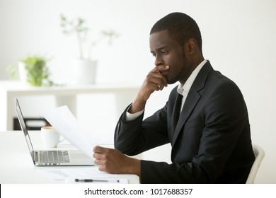 Serious african-american businessman employer thinking of business offer reading mail cover letter at workplace, puzzled black company executive looking at financial document considering contract