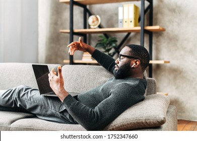 Serious African-American guy in glasses with airpods earphones lays on the sofa in stylish living room and using trendy laptop for video call. Remote work concept