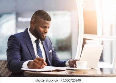 Serious Afro Businessman Working On Laptop Taking Notes During Coffee Break In Outdoor Cafe. Busy Day Concept