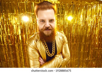 Serious Angry Man Aggressively Looking At You Standing Arms Folded Against Shiny Golden Background. Portrait Of Rude Unfriendly Discontent Rich Guy In Disco Party Jacket And Bling Gold Chain Necklace