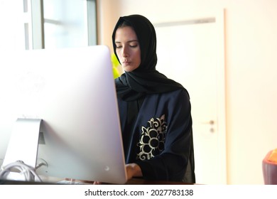Serious Arab woman at office. Working Emirati girl using computer screen wearing traditional Abaya and Hijab Shayla in the Middle East