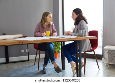 Serious Architects Working And Discussing Design At Desk. Two Women Sitting And Talking In Office With Window In Background. Architects Concept.